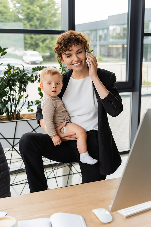 joyful businesswoman holding toddler daughter and talking on smartphone near computer monitor in office