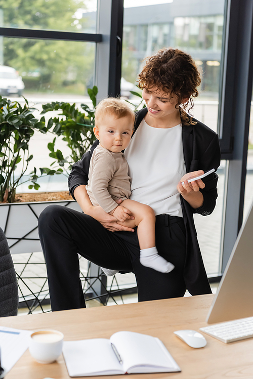 smiling businesswoman with baby and mobile phone near blurred notebook and coffee cup on work desk