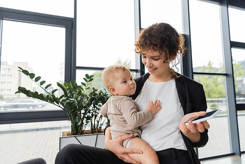 pleased businesswoman holding baby and mobile phone with blank screen near green plants in office