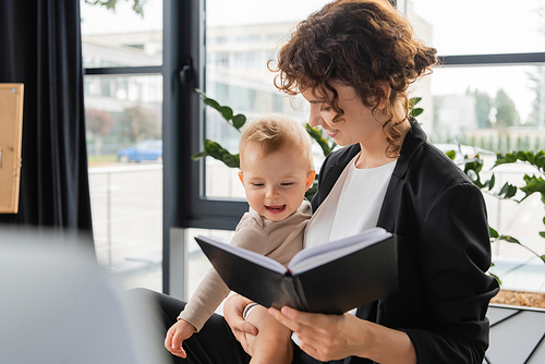businesswoman in black blazer holding notebook and cheerful baby girl in office on blurred foreground