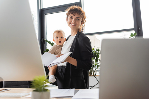 smiling businesswoman holding baby and blank notebook near blurred computers in office