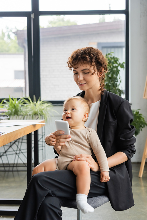 smiling businesswoman in black suit using mobile phone while sitting with toddler daughter in office