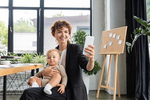 joyful businesswoman in black suit sitting with baby and taking selfie on cellphone in office