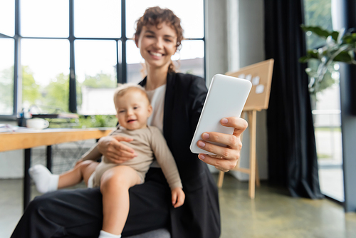 blurred businesswoman taking selfie on mobile phone while sitting with smiling baby in office