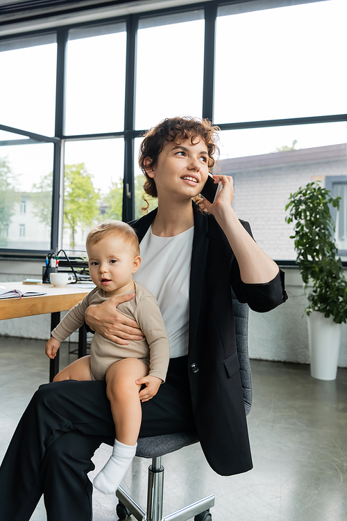 positive businesswoman looking away while sitting with baby in office and talking on smartphone