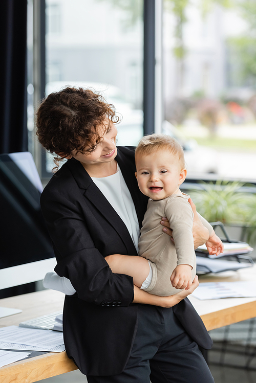 smiling businesswoman standing with little daughter near workplace in office