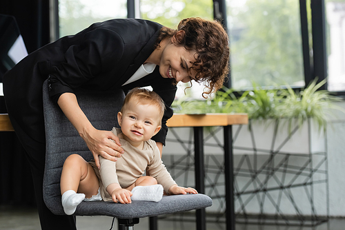 cheerful baby in romper sitting on office chair near smiling mother in formal wear