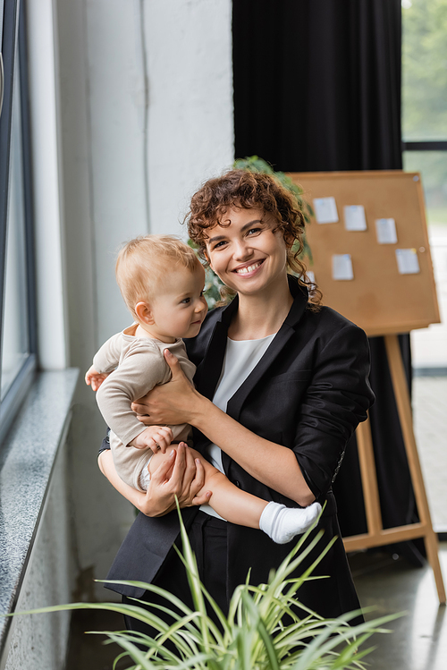 joyful businesswoman in black blazer looking at camera while standing with toddler daughter in office
