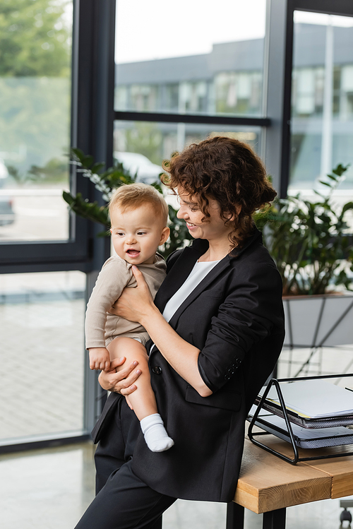 businesswoman in black suit holding little child near work desk with documents in office