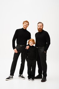 full length of adult bearded men and happy redhead boy standing with hands in pockets and looking at camera on grey background