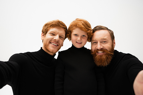 joyful bearded men in black pullovers smiling at camera near red haired boy isolated on grey