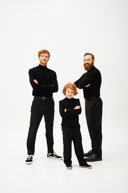 full length of red haired kid with father and bearded granddad in black clothes standing with crossed arms on grey background