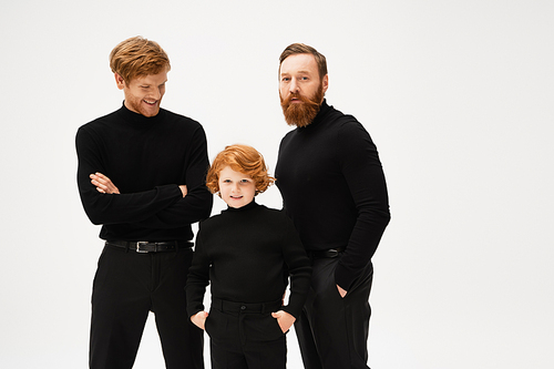 bearded man looking at camera near redhead son and grandson posing in black clothes isolated on grey