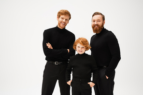 joyful redhead kid standing with hands in pockets near bearded dad and grandfather isolated on grey