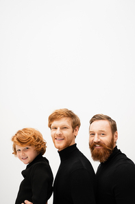 joyful bearded men and redhead boy in black turtlenecks smiling at camera isolated on grey