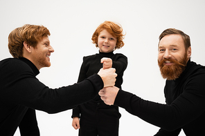 happy bearded men and redhead boy in black turtlenecks joining fists isolated on grey