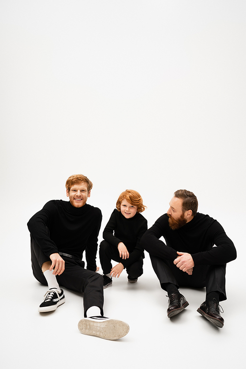 cheerful man smiling at camera while sitting near redhead son and bearded dad on light grey background