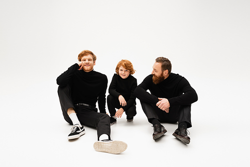 joyful redhead man sitting near smiling son and bearded father in black clothes on light grey background