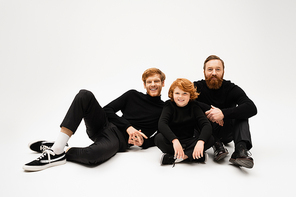 happy bearded men in black clothes smiling at camera near redhead boy while posing on light grey background