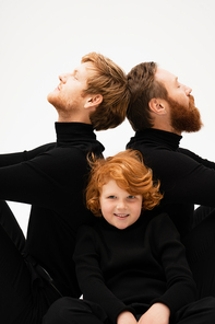 red haired boy smiling at camera near bearded grandpa and father in black turtlenecks sitting back to back isolated on grey