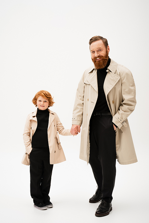 full length of cheerful red haired boy with grandfather holding hands while posing in trench coats on light grey background