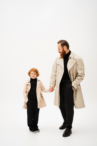 full length of bearded man in trench coat holding hands with redhead grandson posing with hand in pocket on light grey background