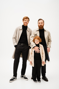full length of smiling red haired boy with father and grandpa in trench coats looking at camera on light grey background