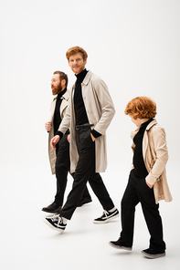 full length of trendy bearded men and redhead boy in trench coats walking on light grey background