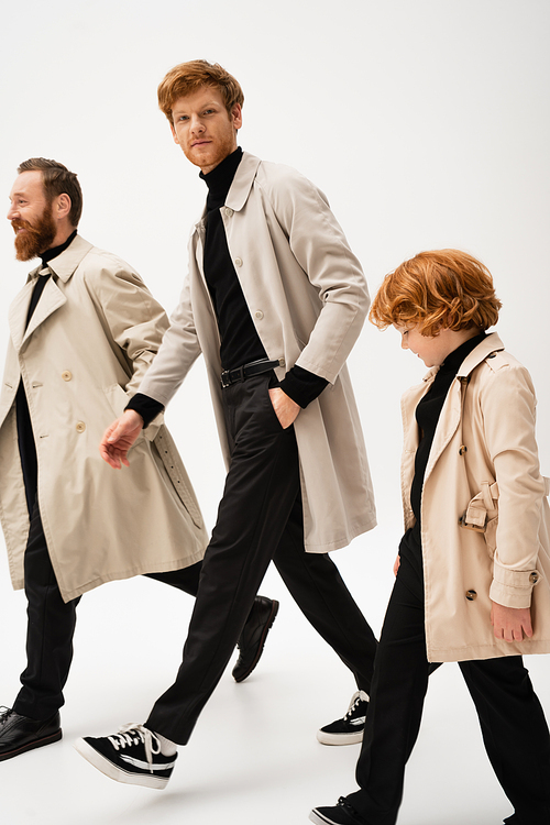 young man in trench coat looking at camera while walking with bearded father and redhead son on light grey background