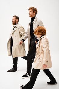 bearded men and redhead boy in trench coats walking on light grey background