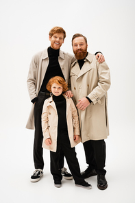 cheerful man hugging bearded dad near redhead son while posing in trench coats on light grey background