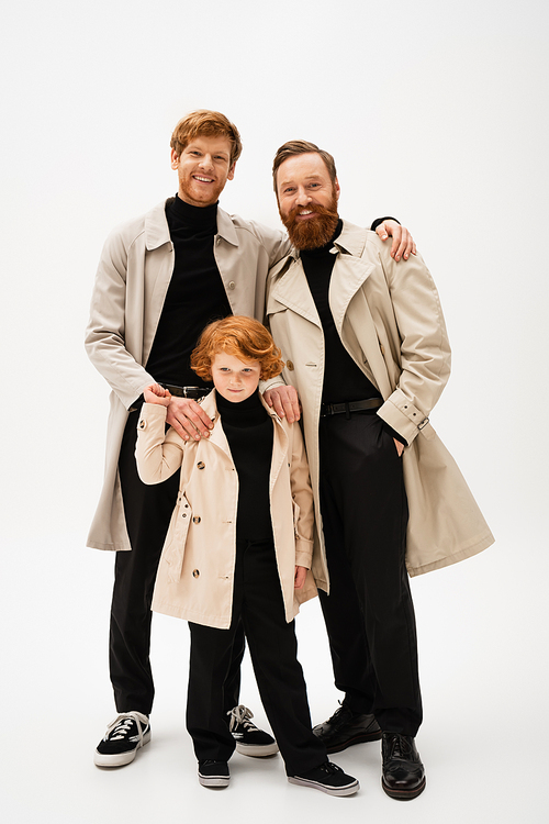 joyful red haired man embracing bearded father and son standing in trench coats on light grey background