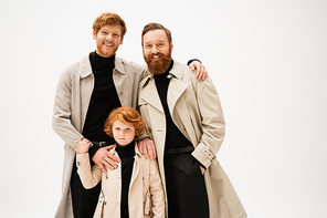 happy bearded men and red haired kid in trench coats looking at camera isolated on grey