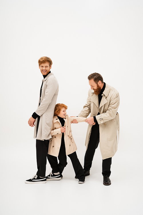 full length of redhead man in trench coat smiling near bearded grandpa and grandson having fun on light grey background