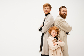 joyful bearded man in trench coats standing back to back with crossed arms near redhead boy isolated on grey