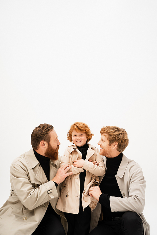 happy redhead kid in trench coat smiling near father and bearded grandpa hugging him isolated on grey