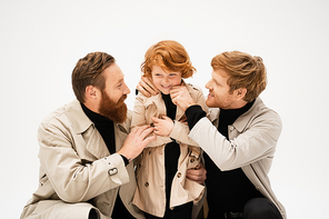 happy bearded men in trench coats embracing cheerful redhead boy isolated on grey