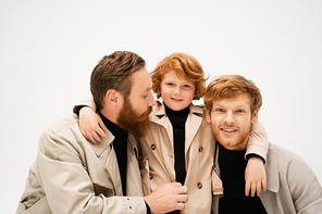 smiling kid in trench coat hugging redhead father and bearded granddad isolated on grey
