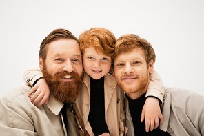 family portrait of red haired boy hugging cheerful bearded grandfather and dad isolated on grey