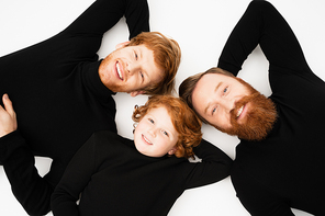 top view of smiling redhead boy with bearded grandfather and dad in black turtlenecks lying on white background