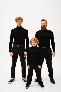 full length of smiling boy and redhead bearded men in black turtlenecks and trousers standing on light grey background