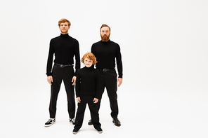 full length of redhead men and kid in black sweaters and trousers standing and looking at camera on light grey background
