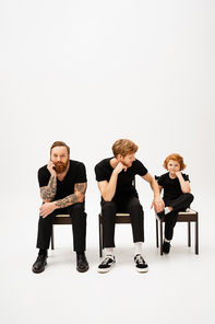 bearded tattooed man smiling at camera near redhead son and grandson sitting on chairs on grey background