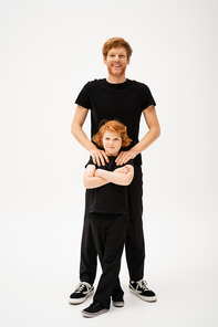 full length of happy man hugging shoulders of red haired son standing with crossed arms on light grey background
