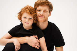 happy redhead man hugging smiling son while looking at camera isolated on grey