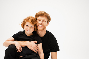 pleased man in black t-shirt embracing red haired kid and looking at camera isolated on grey