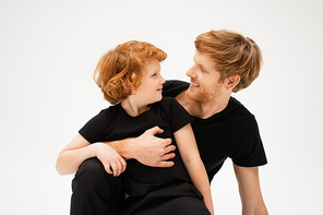 happy man in black t-shirt embracing redhead son while looking at each other isolated on grey