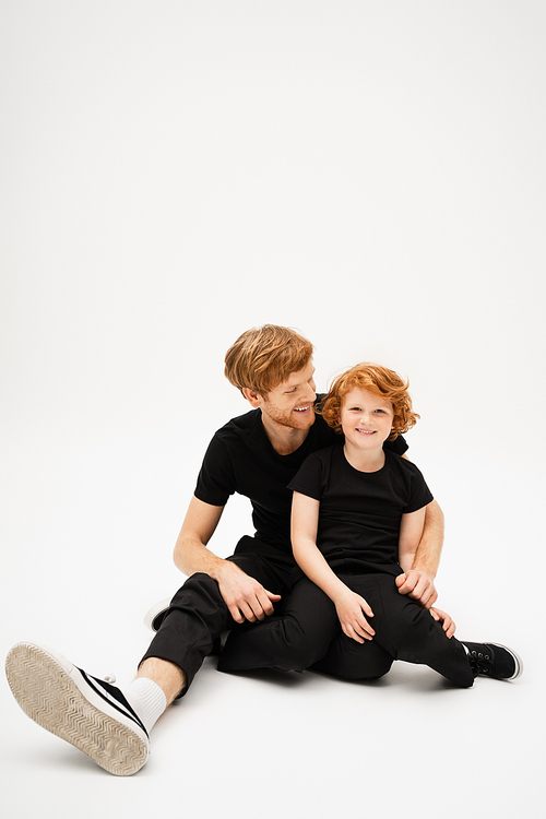 full length of cheerful redhead boy smiling at camera while sitting with young father on light grey background