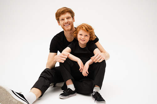 joyful father and son with red hair smiling at camera while sitting on light grey background