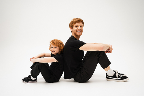 full length of happy red haired dad and son in black t-shirts and pants sitting back to back on light grey background